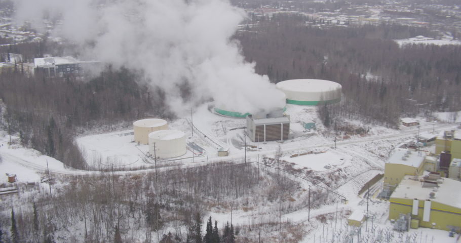 Aerial helicopter shot, tilt down to see steam rising from power plant, cars pass on highway, snow in background, drone footage