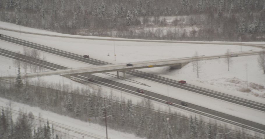 Aerial helicopter shot, cars passing under snowy overpass next to barren forest, drone footage
