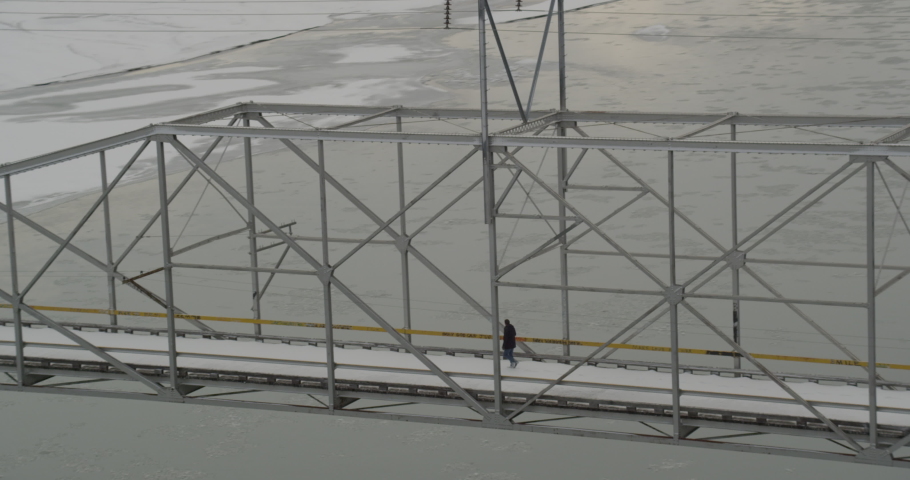 Aerial overhead helicopter shot, circle around track man as walks to center of snowy Alaskan bridge, he looks over both edges at the frozen water, drone footage