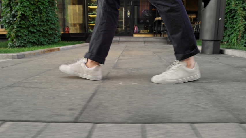 Gimbal shot profile view from ground level of men wearing blue trousers and white sneakers walking along grey pavement on background of grass with sunlight Royalty-Free Stock Footage #1038531398