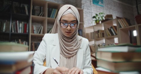 Female muslim student writes down notes in her notebook, sitting at desk covered with books and looking at camera - modern islam, student lifestyle concept close up 4k portrait