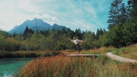 Young female is doing yoga on her mat in beautiful nature at a mountain lake. She contemplates a Lord of the Dance Pose. Yoga and meditation have good benefits for health.: stockvideo