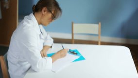 Mature serious female doctor sits at a table in the office and writes out a prescription for the patient. Healthcare and medicine concept, dynamic video