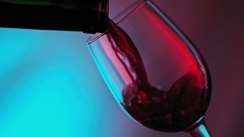 Red wine forms beautiful wave. Wine pouring in wine glass at red and blue background. Slow motion of pouring red wine from bottle into glass. Close-up shot.
