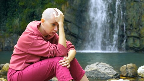 Sad bald woman sits on a stone against the background of a waterfall. copy space. concept of unity with nature, hiding from people.
