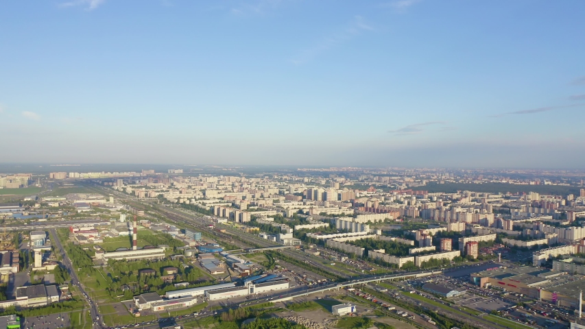 St-Petersburg, Russia. Aerial view over the Parnas district revealing a sweeping panorama of modern residential blocks, mid-rise apartment buildings, and scattered high-rise towers spreading across the northern cityscape. The district takes its name from an artificial hill commissioned by the noble Shuvalov family in the late 18th century, referencing the Greek Mount Parnassus. The Severnaya Dolina housing complex, built across multiple phases in the post-Soviet era, anchors the area with connected residential massifs and towers reaching up to 25 floors. A wide arterial highway cuts through the foreground carrying steady traffic, while patches of green parkland and tree-lined courtyards punctuate the dense urban fabric. The scene is bathed in warm late-afternoon sunlight under a clear pale-blue sky, casting soft golden tones across rooftops and facades. The camera drifts smoothly above the city, offering a broad top-down perspective that emphasizes the scale and geometric layout of this contemporary residential zone on the edge of the historic metropolis.