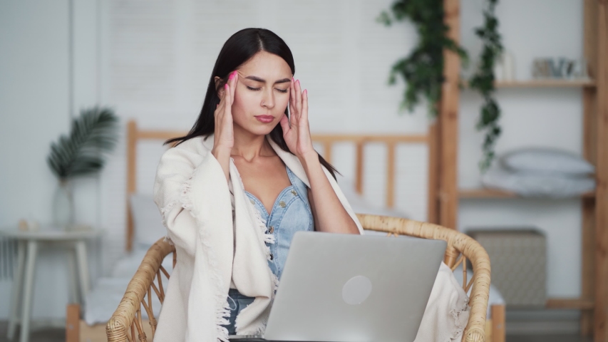Portrait of tired woman works on laptop at home and has horrible headache. Girl touches her head