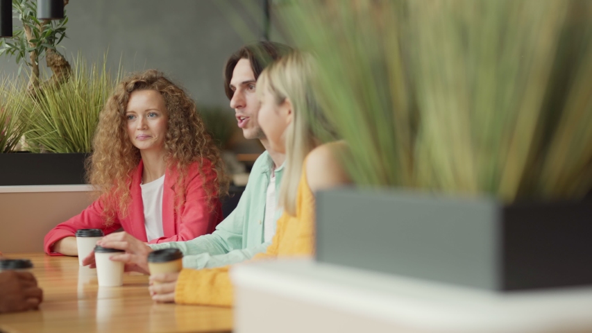 Camera panning left to young man gesturing and telling funny story to laughing multiethnic friends while sitting at table with coffee to go in cafe