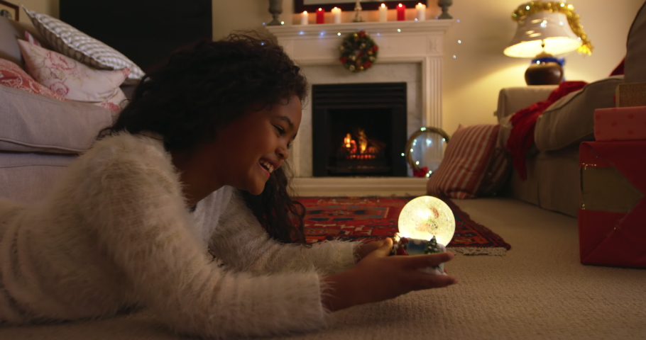 Side view of a young mixed race girl in her sitting room at Christmas lying on the floor, smiling and looking at a snow globe