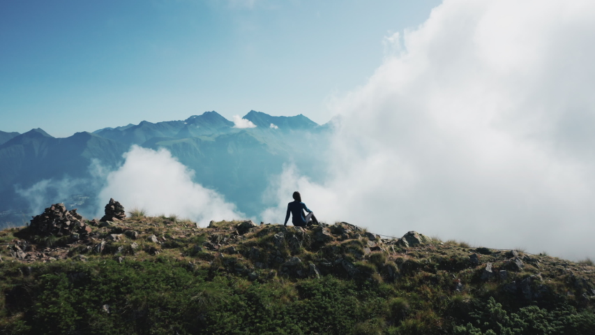 Young brave girl seating on cliff edge enjoying panorama, feeling fresh air; thick clouds moving around; woman silhouette against the mountains; sunny autumn day, aerial view, drone flying forward Royalty-Free Stock Footage #1038592433