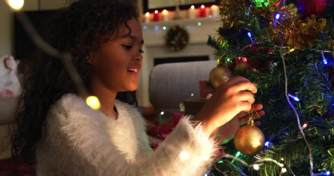 Side view close up of a young mixed race girl in her sitting room at Christmas decorating the Christmas tree and smiling with joy, her father visible sitting in the background Arkivvideo