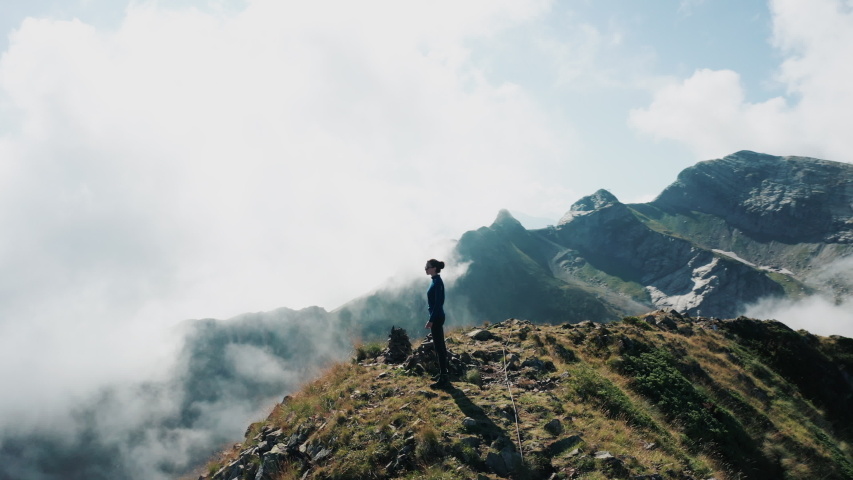 Young brave girl standing on cliff edge enjoying panorama, feeling fresh air; clouds moving around; woman silhouette against mountains; freedom and independence; sunny summer day; aerial drone view Royalty-Free Stock Footage #1038593759