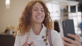 Close up portrait of Caucasian woman with curly hair standing in store with paper shopping bags in her hands and having online video talk