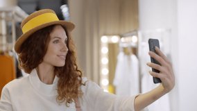 Close up of joyful woman with curly hair standing in apparel shop with smartphone in her hands and hat on her head and making selfie with smile on her face