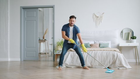 Slow motion of attractive young man in rubber gloves washing wooden floor and dancing with plastic mop having fun in flat. People and housework concept.