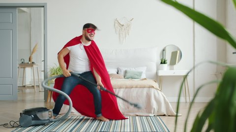 Crazy guy in super hero costume is dancing with vacuum cleaner having fun indoors vacuuming carpet in bedroom. Funny people, super hero and lifestyle concept.