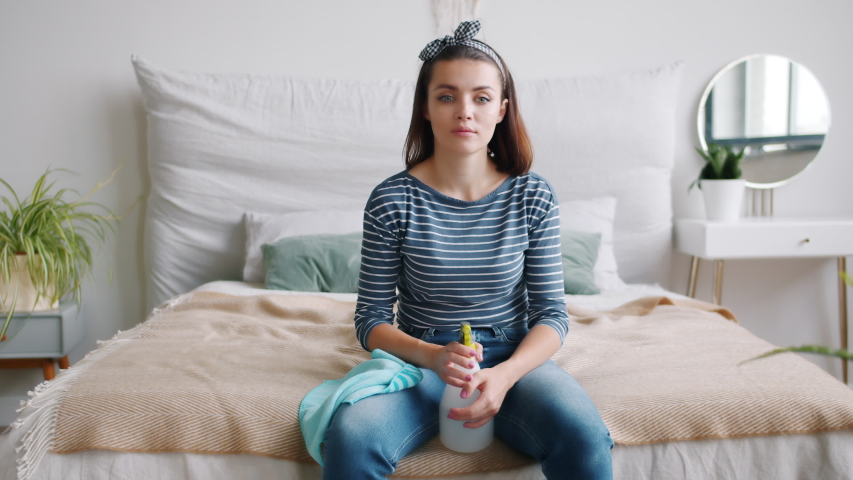 Tired housewife attractive young woman is sprinkling water on face and hands from spray bottle during clean-up sitting on bed in apartment. People and housework concept.