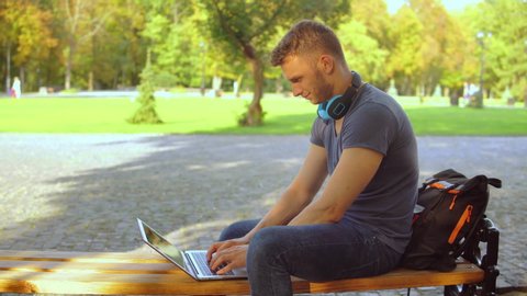 handsome young man typing on laptop sitting on the bench working in city park. autumn season outdoors. beautiful view on nature green and yellow colors