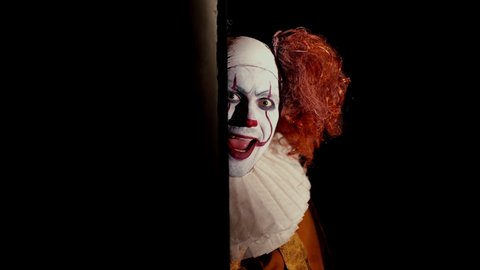 Crazy clown with a wide, frightening smile slowly appears from a dark corner. Shooting in black room, close-up face