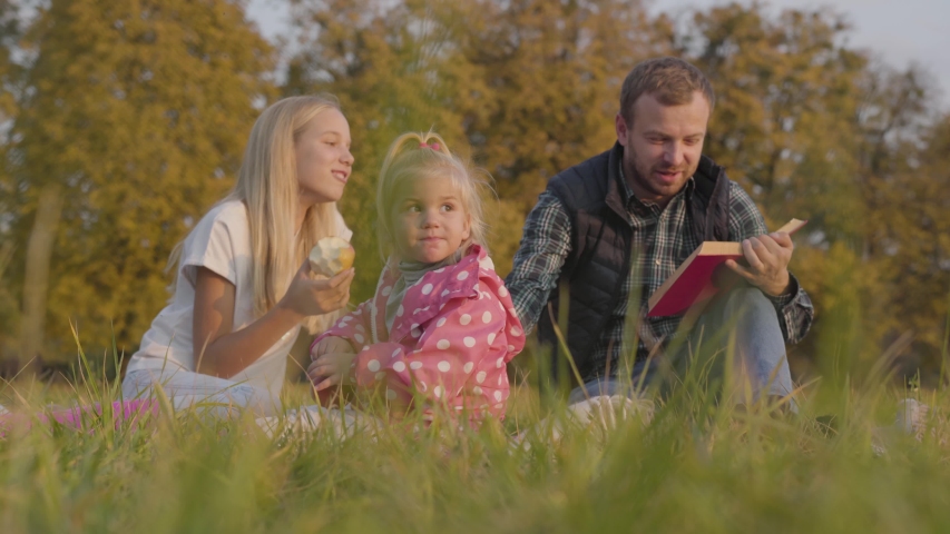 Happy family sitting on the autumn meadow, eating apples and reading the book in red cover. Bearded caucasian man having picnic with two blonde pretty girls outdoors.