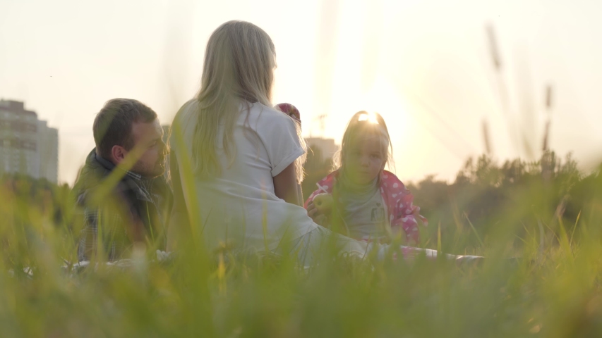 Happy caucasian family sitting on the meadow in sunrays and eating apples. Father spending time with his teen daughter and little blonde girl outdoors.