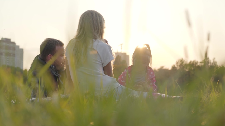 Two caucasian girls and their father sitting on the meadow in sunrays and eating apples. Happy family spending time together outdoors.