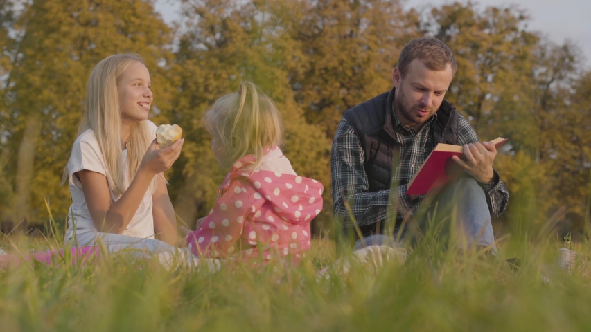 Bearded caucasian man reding book in red cover to his adorable daughters outdoors. Pretty teenager and blonde little girl eating apples on the autumn meadow and listening to their father.