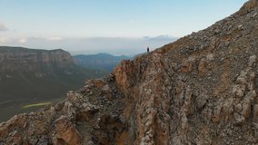 Aerial view of a girl traveler with a backpack stands on the edge of a cliff of a high rocky mountain and looks at the snow-capped mountains in the distance. Travel video drone footage