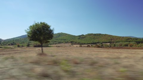 Rural scenery speeding car POV side window day sunny view motion blur. Point Of View looking through car open window, driving on empty road country side with green hills and plantation in Greece.