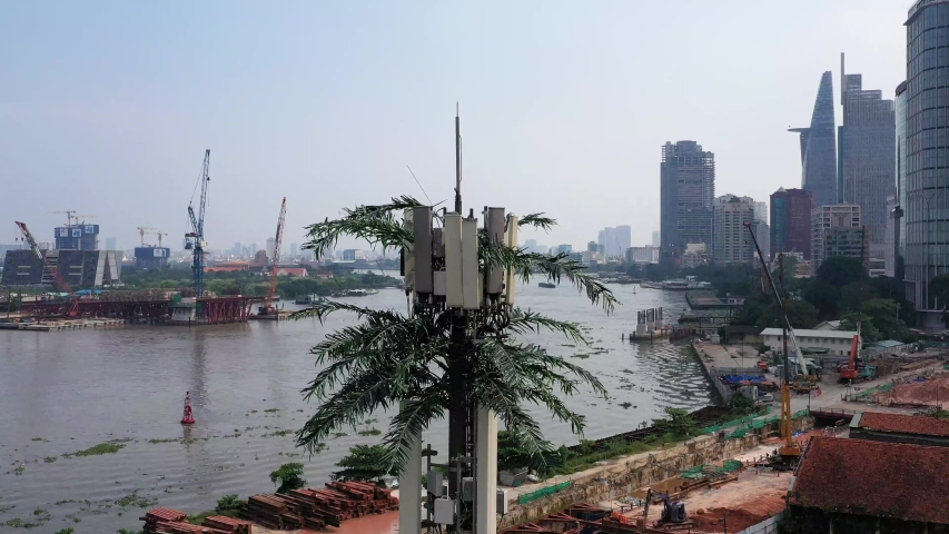 descending aerial shot of ultra modern mobile phone tower disguised as a palm tree against a background of a riverfront construction site with city buildings, cranes and boat traffic on the river - Powered by Shutterstock - Get 15% off with code: PIKWIZARD15