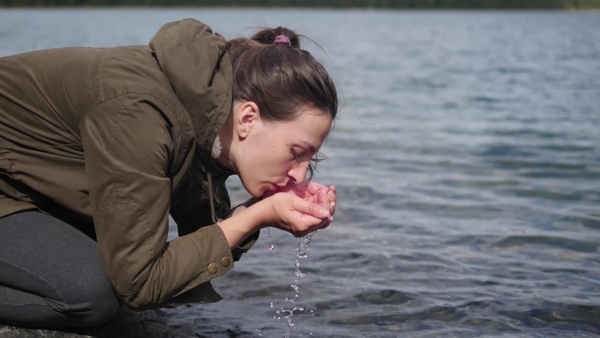 A woman drinks crystal clear water from a lake.