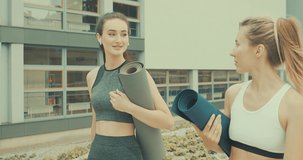Beautiful young girls  talking, go to sports training with yoga mat, in city park in summer sunny day under sun light. 4K slow motion video
