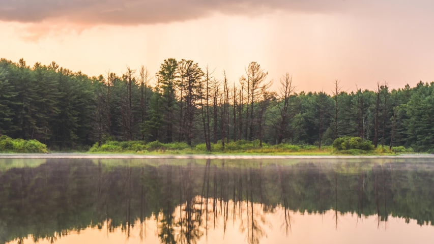 Sunset timelapse of forest treeline reflecting on pond with heavy clouds moving above and a gentle fog on the water.