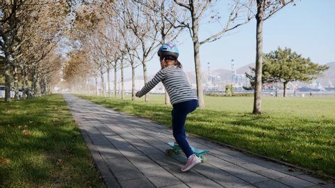Little girl is slowly skateboarding in protective helmet in the park on path at sunny autumn weather, steadicam shot in slow motion.