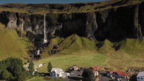 A small Icelandic rural farm lies in front of a long, impressive waterfall and mountain cliff. South Iceland, Europe