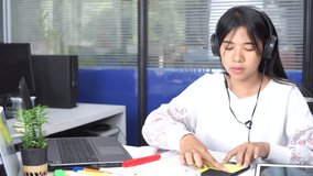 Asian woman freelancer talking writing for planning and video conference online with headphones and laptop in office computer room or home desk with happy for work, Custom support business concept