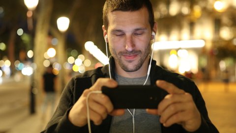 Happy adult man wearing earbuds watching online videos on mobile phone walking towards camera in the street in the night