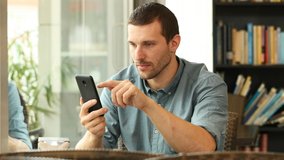 Worried man complaining checking smart phone content sitting in a coffee shop