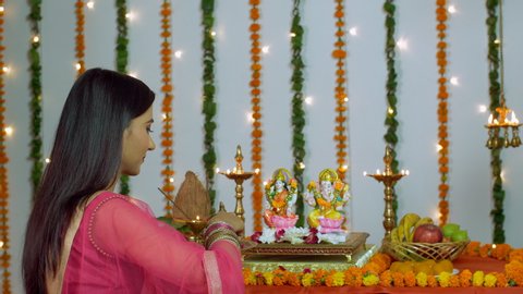 Beautiful Indian woman praying in front of Hindu gods Laxmi and Ganesh - Diwali/Dipavali. Young female offering flowers in the decorated temple of Lord Ganesha and Goddess Lakshmi while doing Diwali...