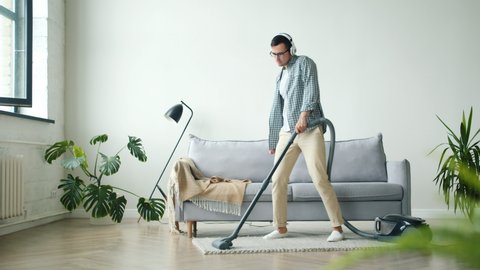 Young guy student is cleaning carpet with vacuum cleaner listening to music through headphones dancing having fun alone at home. People and household concept.
