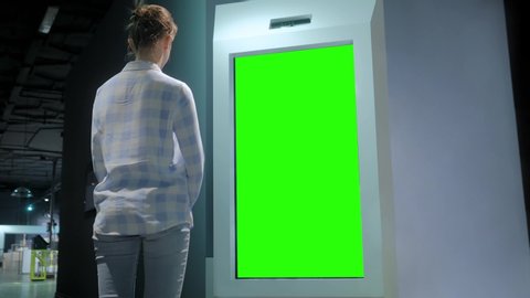 Green screen, mock up, future, copyspace, template, chroma key, technology concept. Woman looking at blank interactive touchscreen green display of electronic kiosk at futuristic exhibition or museum