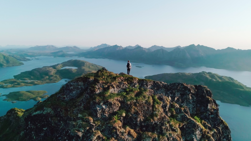 4K drone shot with orbit motion of Norwegian climber standing on the top of tall mountain looking at and admiring the panorama sunset background view of Lofoten and Vesterålen in Norway. Royalty-Free Stock Footage #1038701126