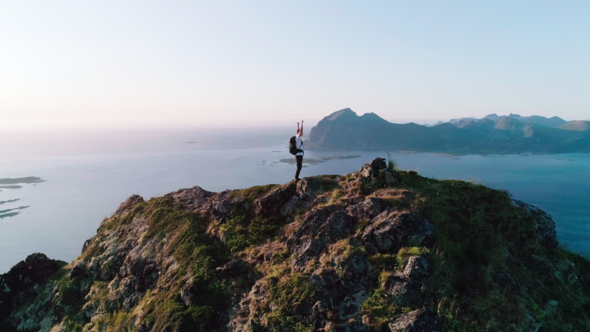 4K drone shot with orbit motion of Norwegian climber celebrating climbing top of mountain by raising his arms to the sky with panorama sunset background view of Lofoten and Vesterålen, Norway. Royalty-Free Stock Footage #1038701135