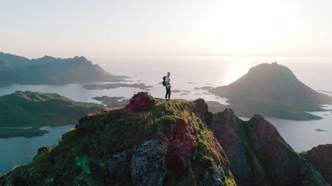 4K drone shot with orbit motion of Norwegian climber celebrating climbing top of a mountain by raising his arms to the sky with panorama sunset background view of Lofoten and Vesterålen, Norway.