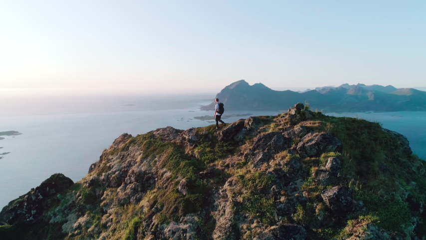 4K drone shot with orbit motion of Norwegian climber celebrating climbing top of a mountain by raising his arms to the sky with panorama sunset background view of Lofoten and Vesterålen, Norway. Royalty-Free Stock Footage #1038703265