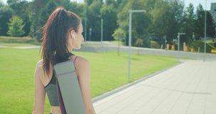 Beautiful young woman go to sports training with yoga mat and listening music in headphones in smartphone at city walk in summer sunny day under sun light. 4K slow motion video
