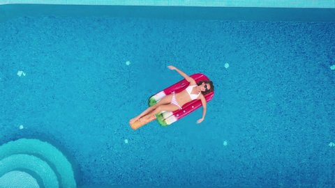 Sexy woman rest and sunbath on a float in the pool, top view aerial shot. Young woman in a bikini swimsuit floating on an inflatable pink mattress top view