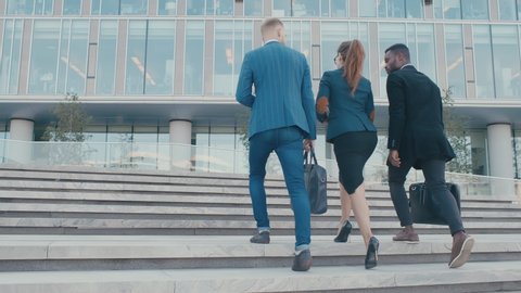 Confident team members walking on stairs in slow motion. Business men and woman in formal suits go and talk on the background of modern office building. Concept of successful employees white collars