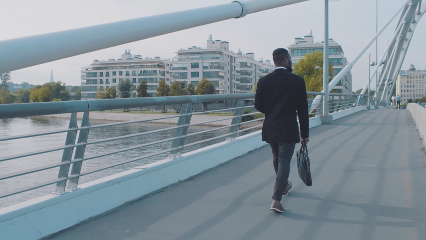 Confident young african american businessman with briefcase walking on modern pedestrian bridge in rich disctrict of city outdoors. Bearded black man on background of modern buildings. Shot from back