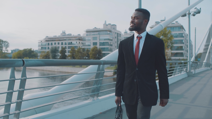 Confident young african american smiling businessman with briefcase walking on modern pedestrian bridge in rich disctrict of city outdoors. Happy bearded black man on background of modern buildings